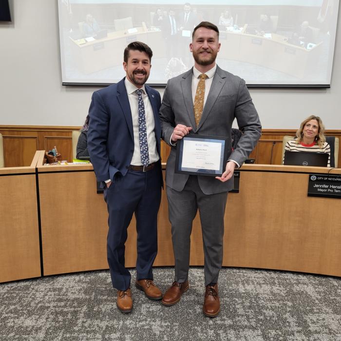 Two people stand in a meeting room, one holds a certificate and both are smiling.