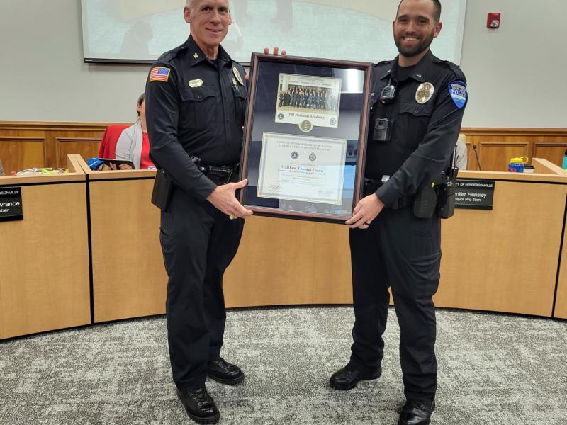 Two police officers stand in a conference room, holding a framed certificate.