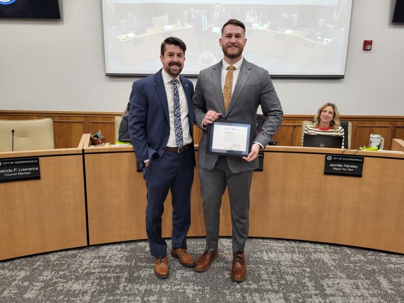 Two people stand in a meeting room, one holds a certificate and both are smiling.