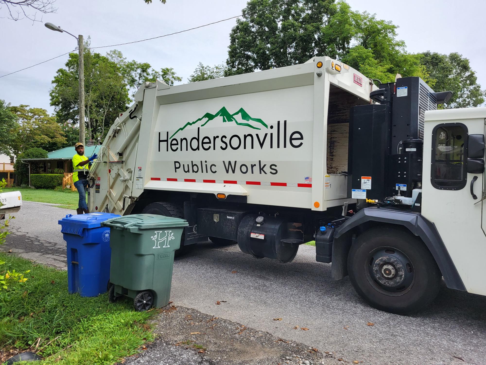 Garbage truck parked with a worker on the back about to pick up a recycling and trash can