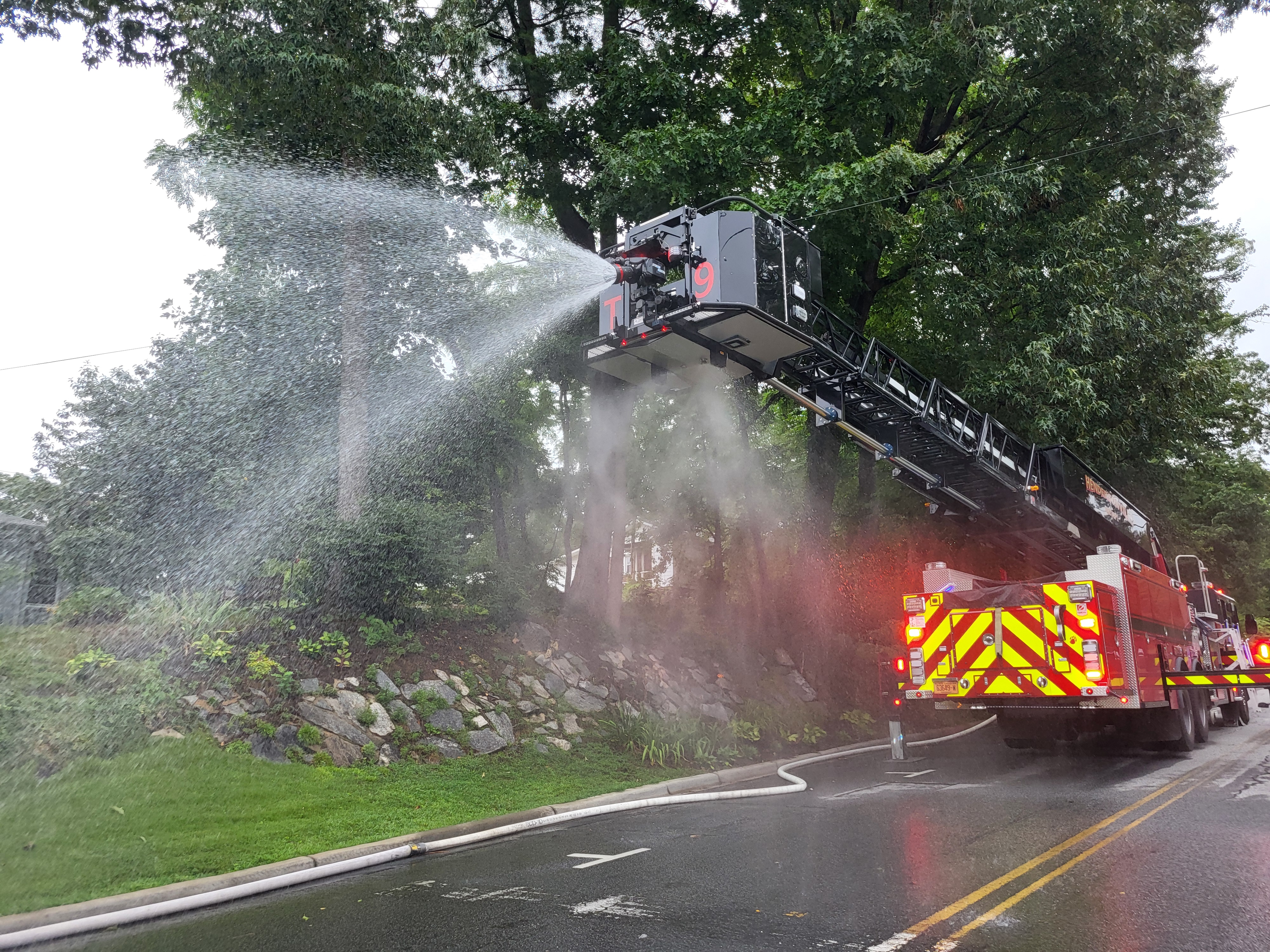 Red fire truck with the ladder raised, spraying water across a closed street.