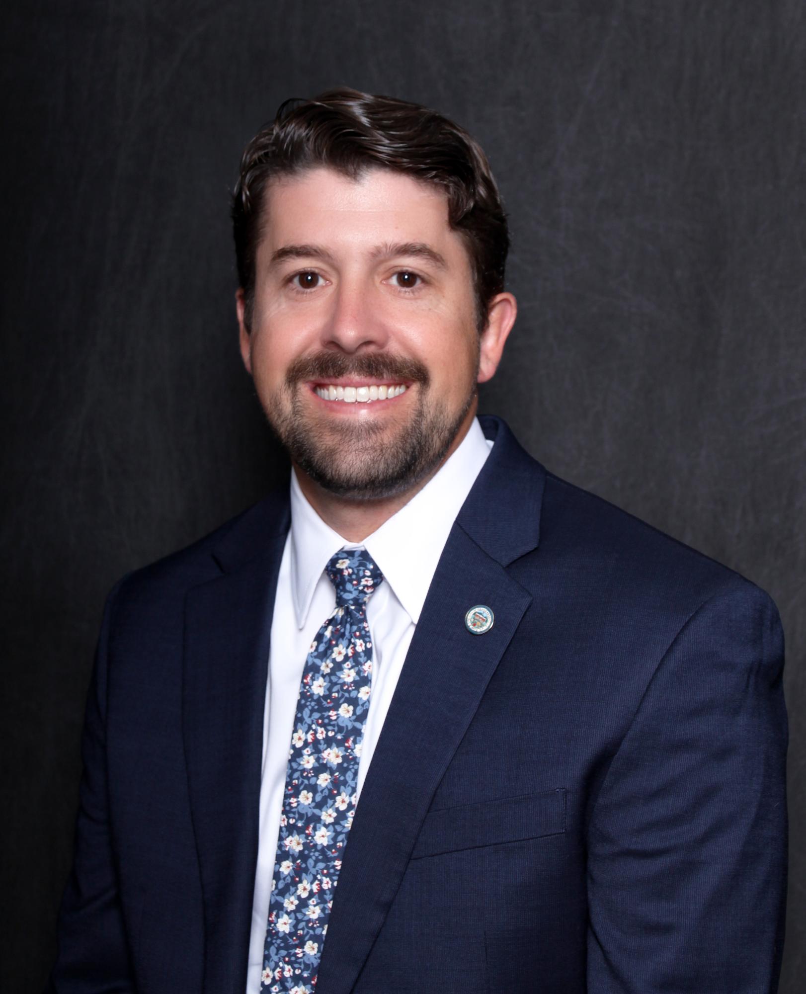 Man in a dark blue suit with a light blue tie is smiling.