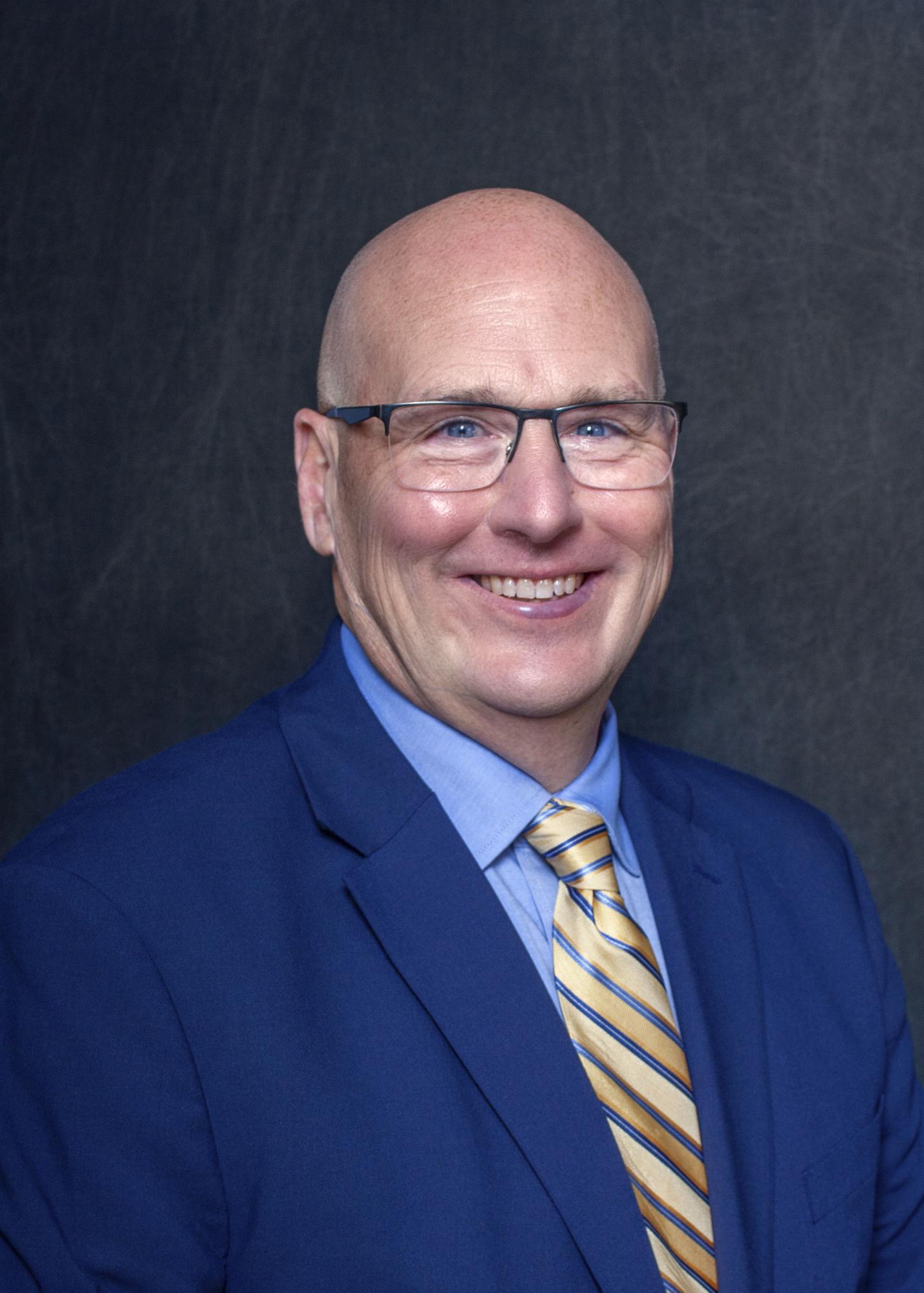 Headshot of a man wearing a dark blue suit, yellow tie and glasses, and smiling.